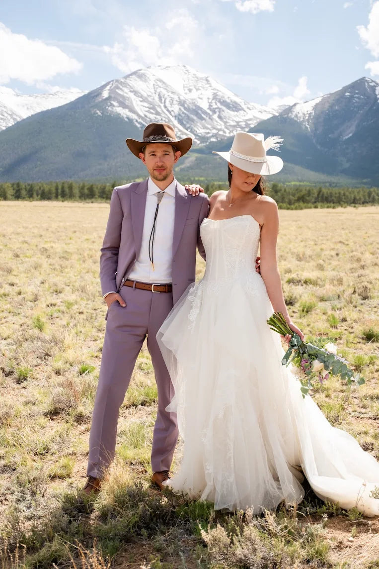 Couple in wedding attire and bolo tie standing in a field with mountains in the background