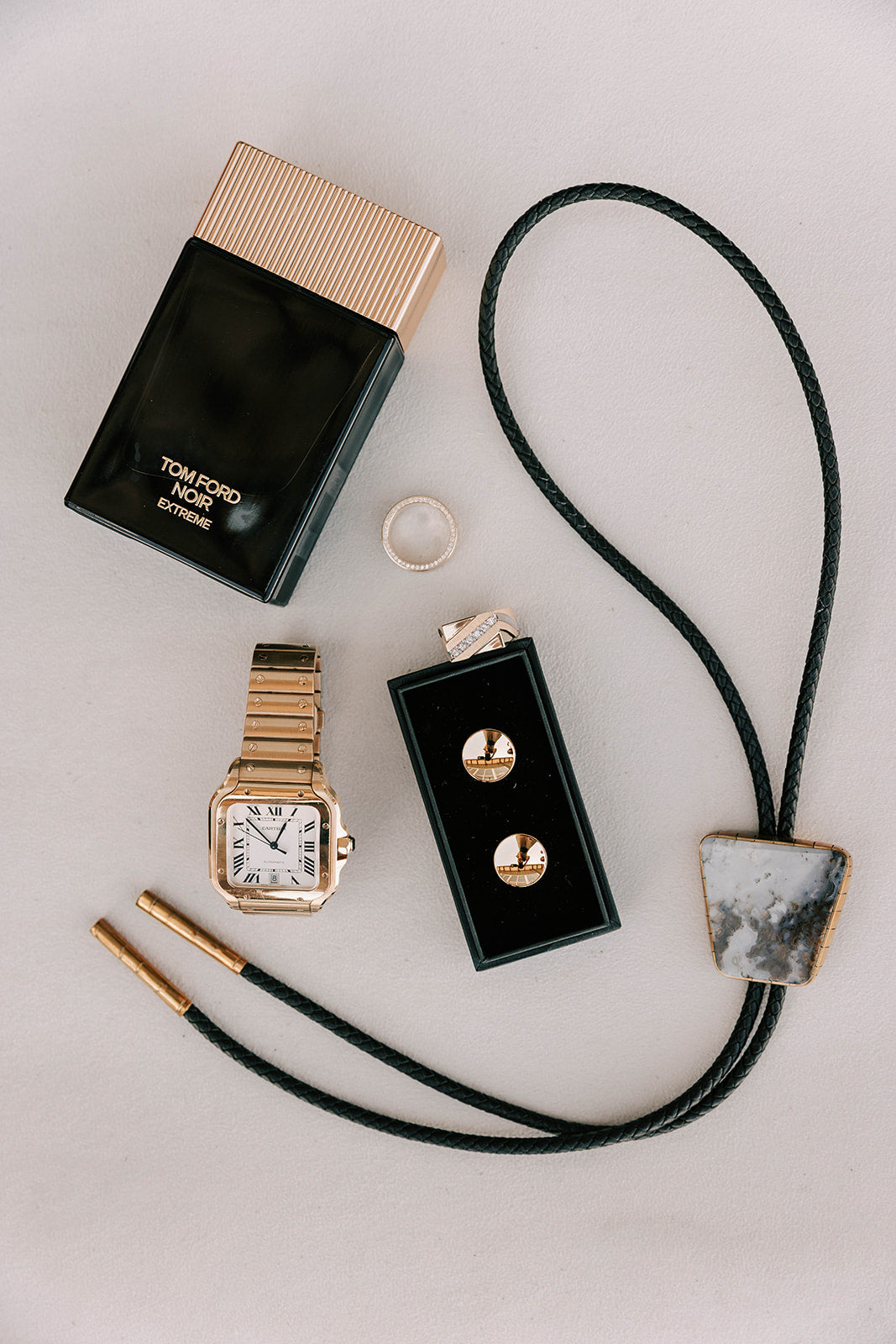 Tom Ford perfume bottle, gold watch, graveyard point bolo tie, and earrings on a light gray background